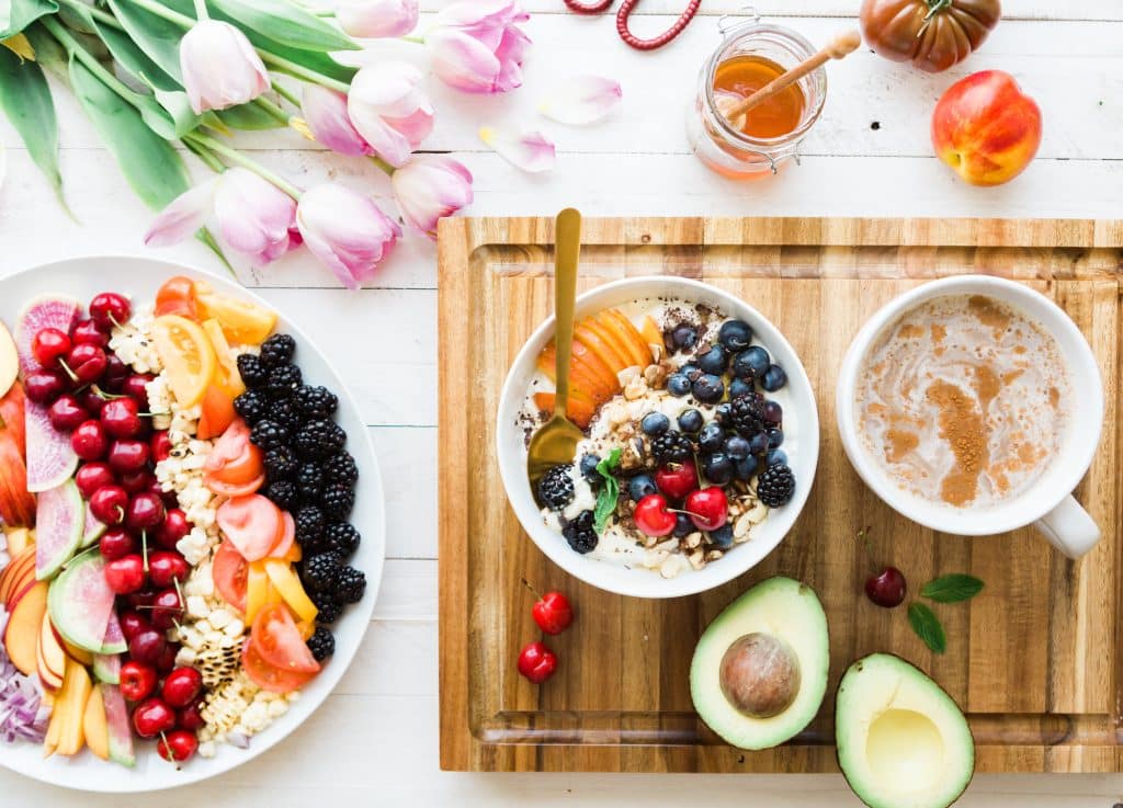 A sliced open avocado, healthy cereal with berries, and other healthy diet foods laid out with honey and flowers