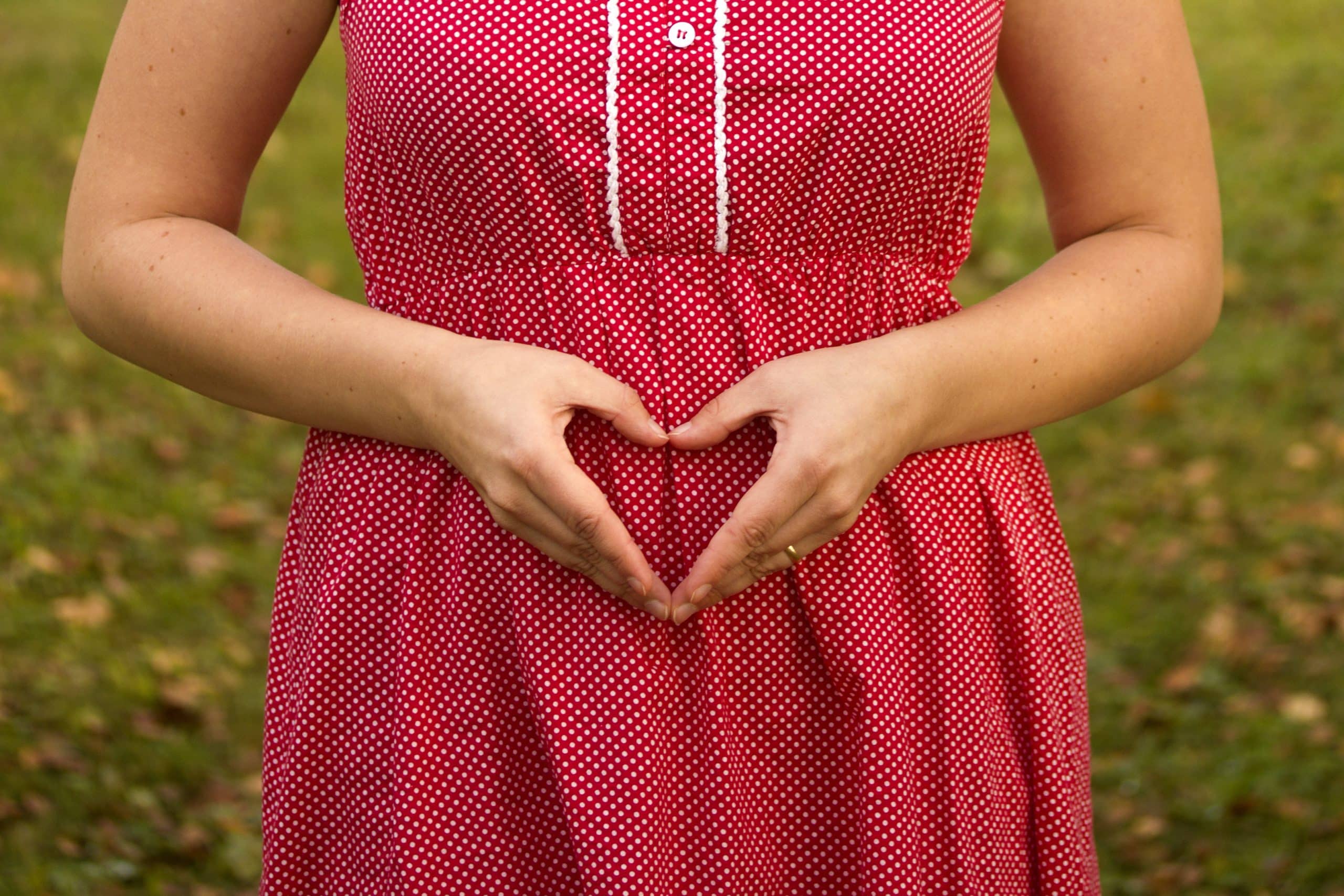 Woman wearing a red polka dot dress forming a heart over her belly with her hands as a sign of pregnancy