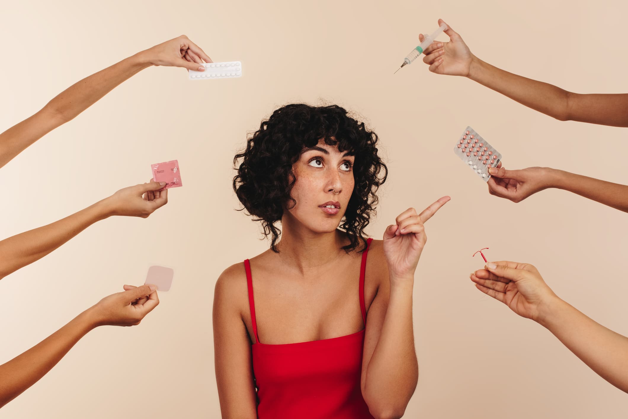 Young woman surrounded by hands holding different birth control methods and pointing at her choice