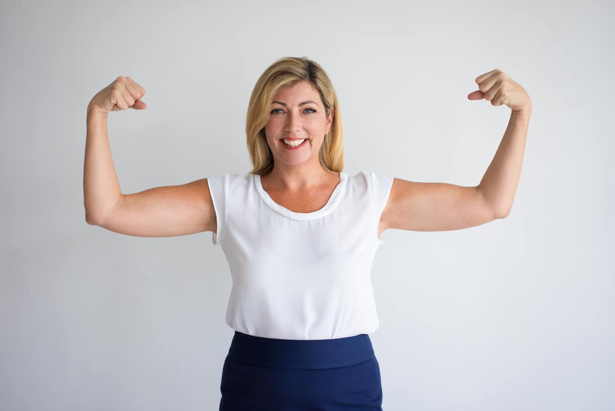 Happy blonde woman flexing biceps in a white shirt and navy pants.