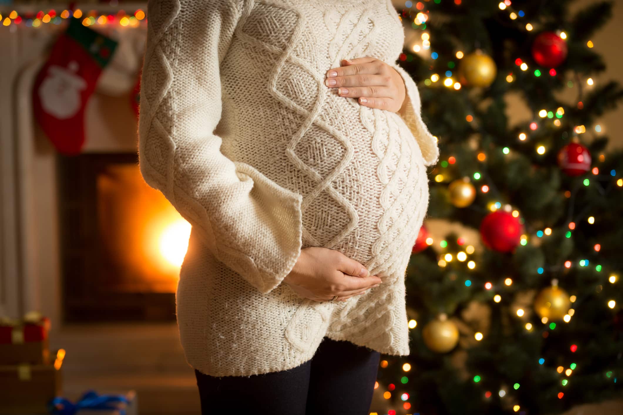 A pregnant woman in a white sweater holding her stomach in front of a Christmas tree and fireplace.