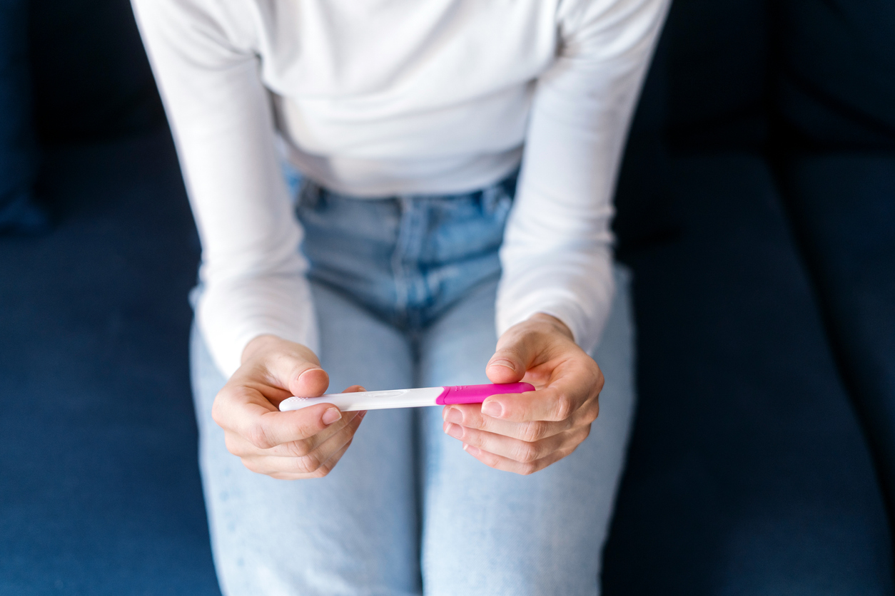 A woman in jeans and a white shirt sitting on a couch gazing at a pregnancy test.