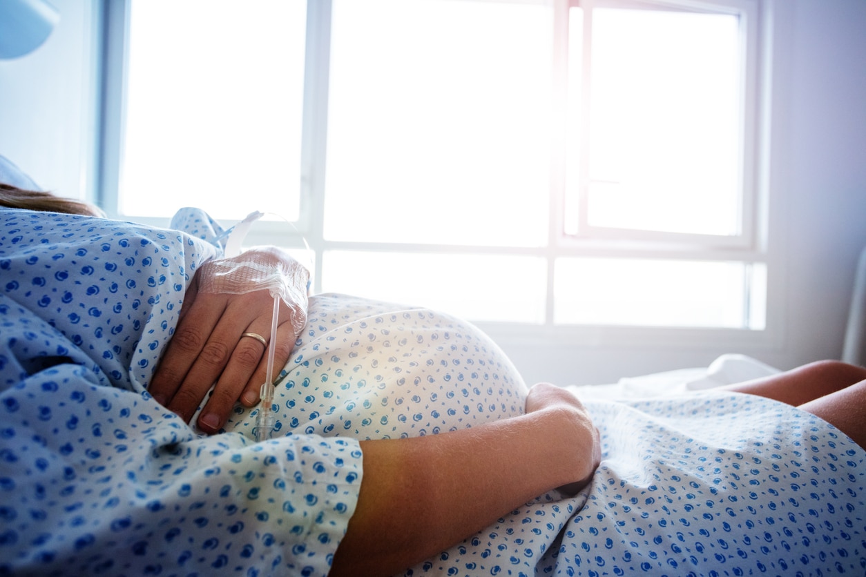 A pregnant woman in labor wearing a hospital gown and resting her hand on her belly in a sunny hospital room.